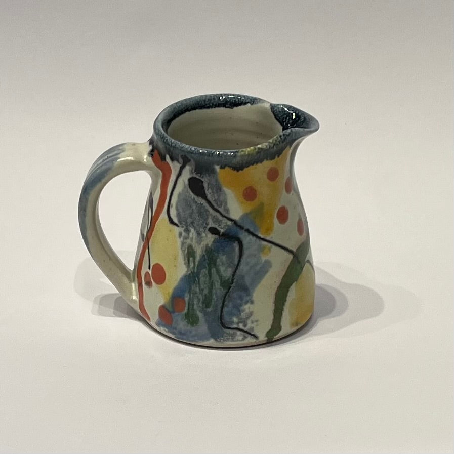 A small ceramic jug, decorated with abstract brushstrokes and splatters in blue, yellow, green, orange, and red on a white background.